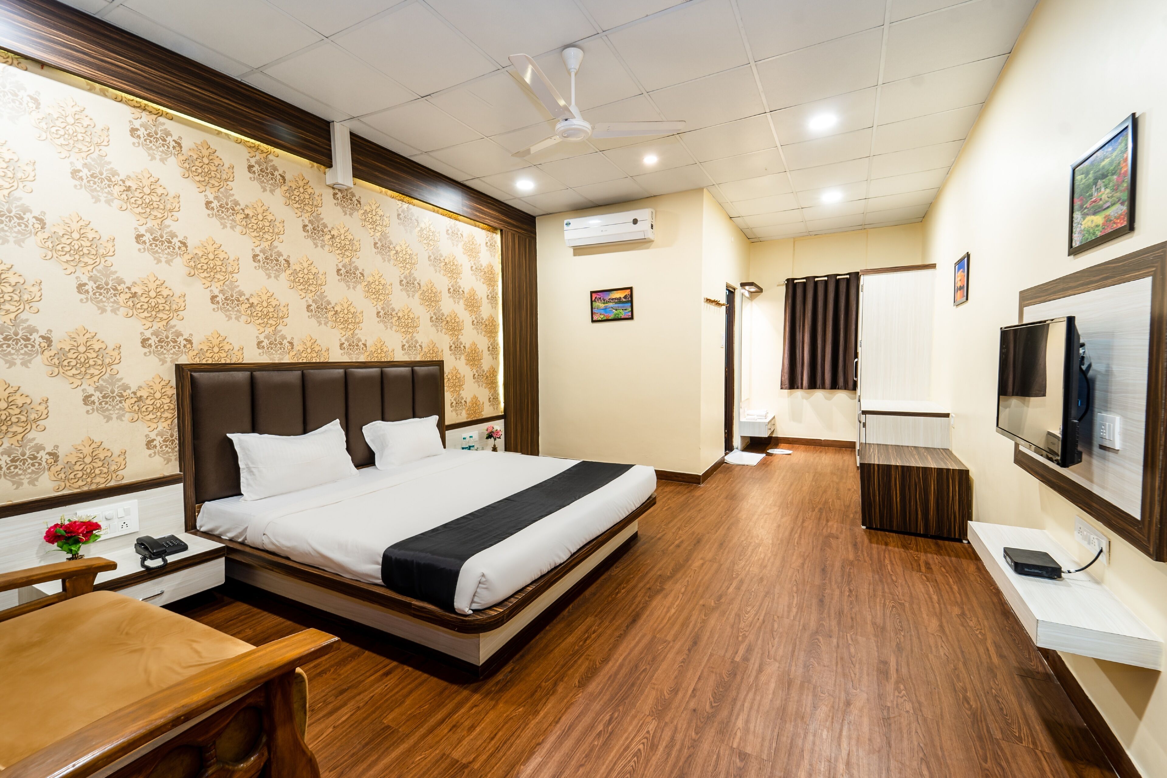 A hotel room with a large bed, wooden flooring, a desk, a TV, and an air conditioning unit.