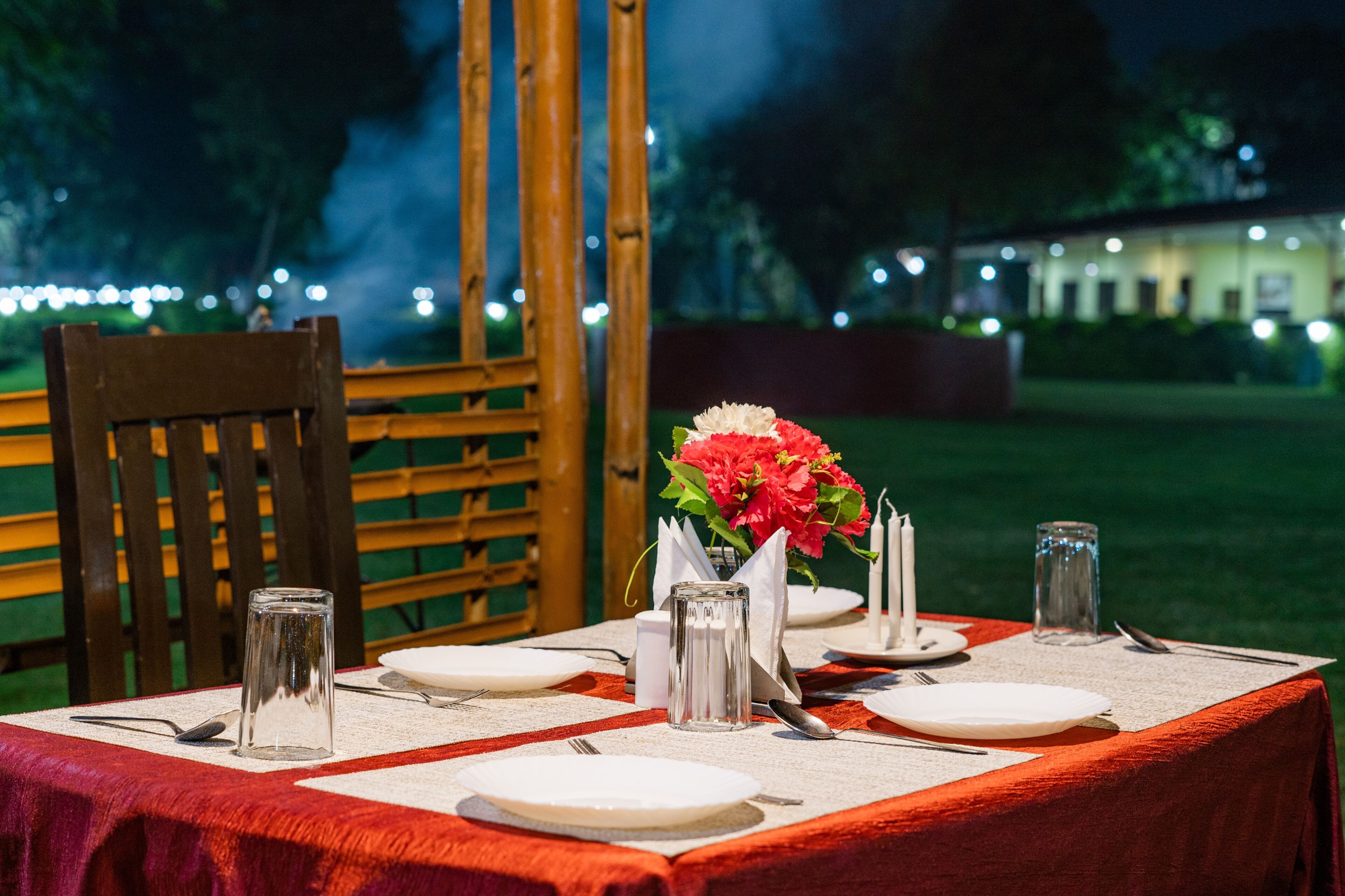A table set for dining outdoors at night with a red tablecloth, white plates, cutlery, and a vase with flowers.