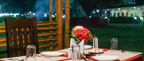 A table set for dining outdoors at night with a red tablecloth, white plates, cutlery, and a vase with flowers.