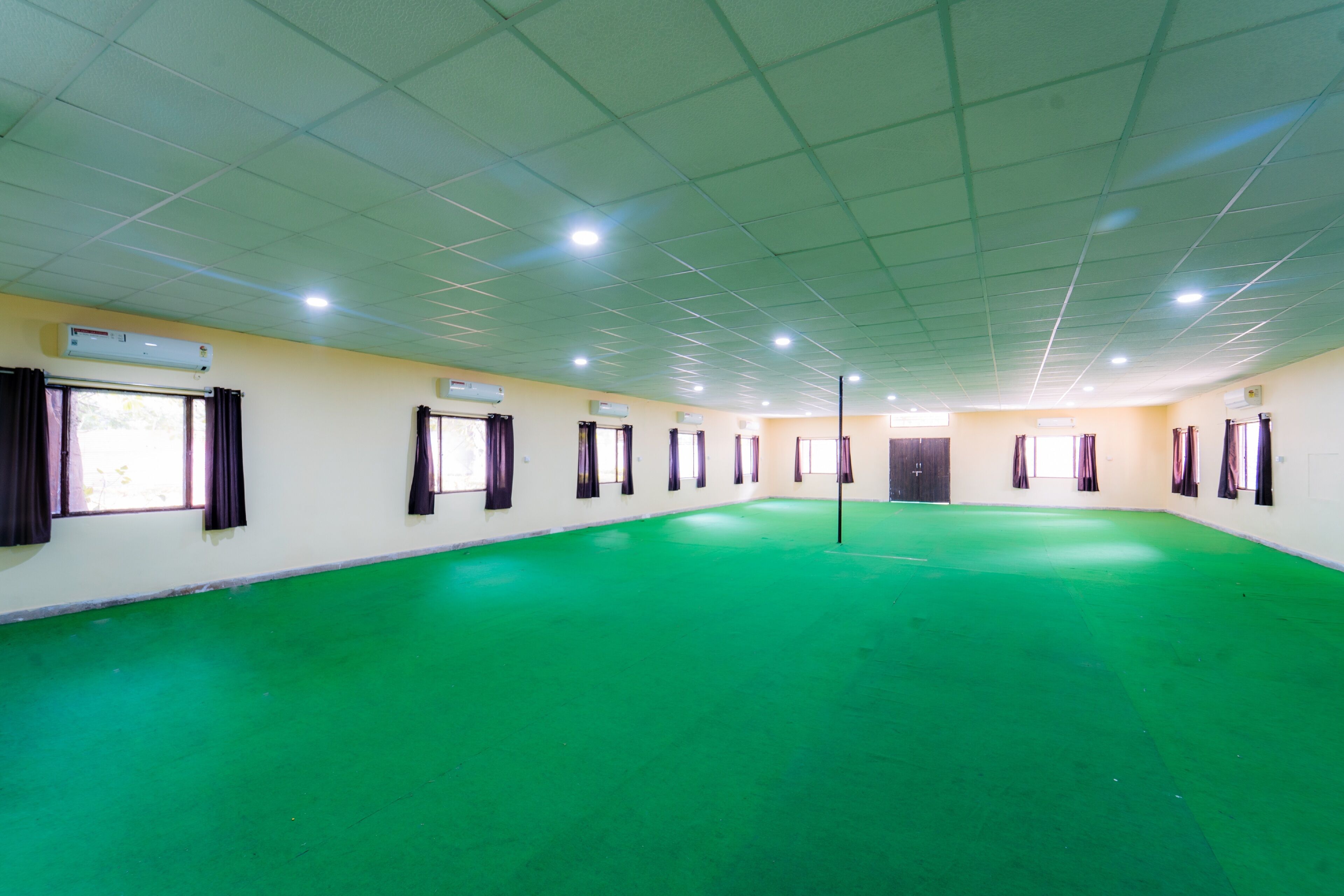 A large, empty room with green flooring, white walls, and multiple windows with dark curtains.