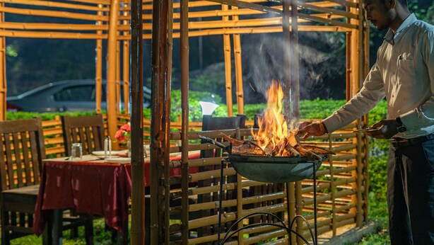 Una persona cocinando al aire libre usando una parrilla, una mesa con mantel rojo y sillas.