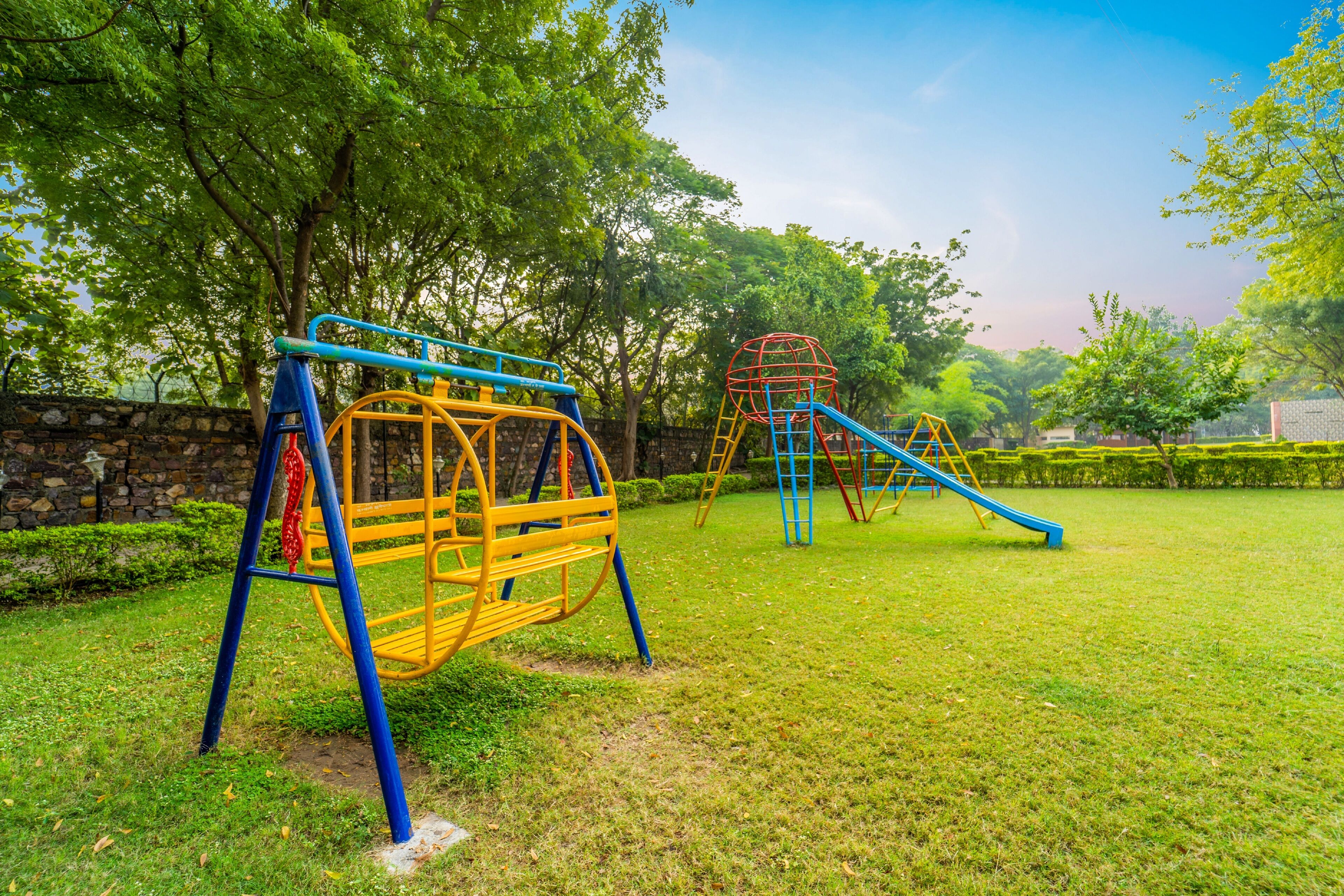 A playground with swings and slides, surrounded by trees and a stone wall.