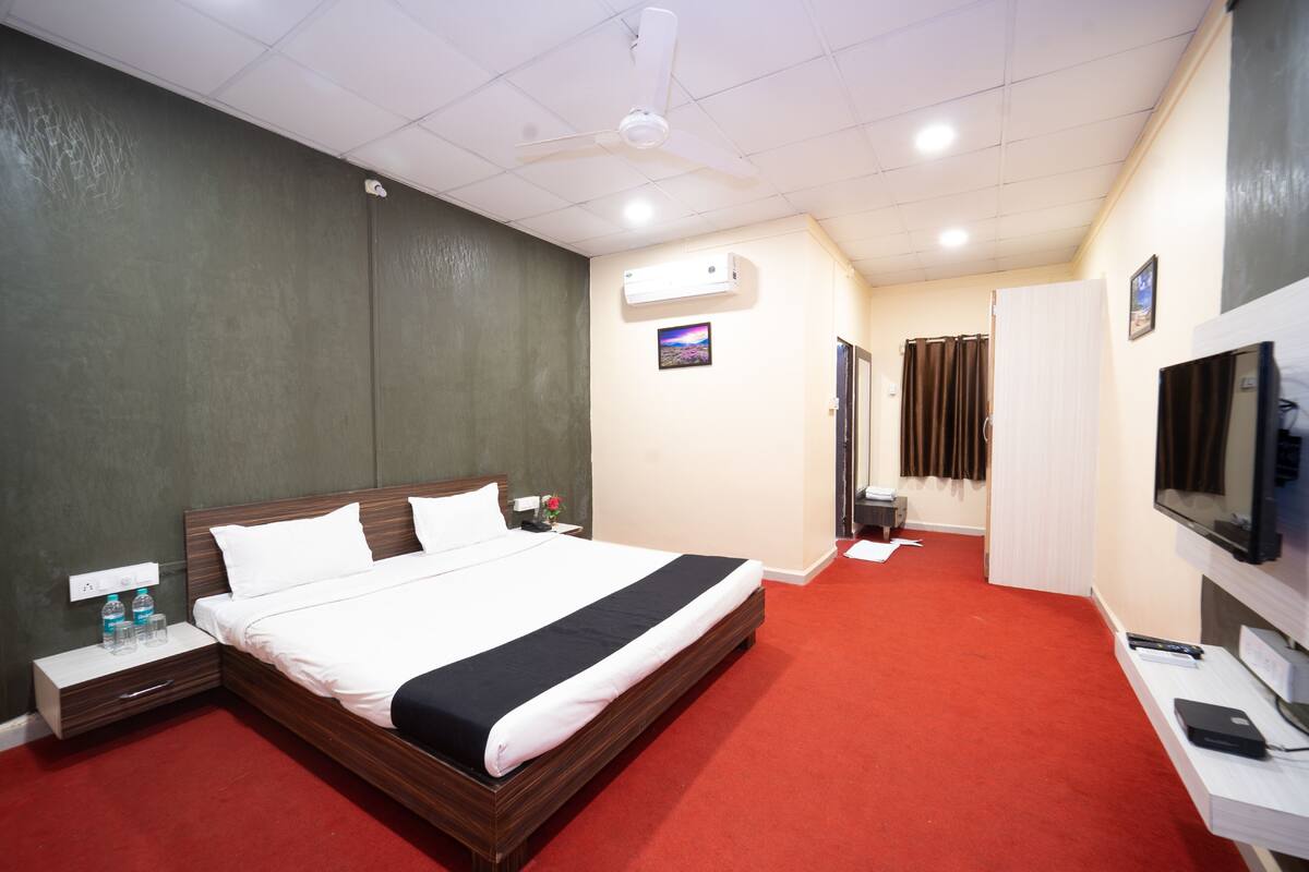 A hotel room with a large bed, a desk with a computer, a TV, and an air conditioning unit on the wall.