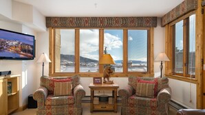 Ein gemütliches Wohnzimmer mit einem großen Fenster, zwei gemusterten Sessel, einem kleinen Tisch mit Lampe sowie einem Fernseher an der Wand.
