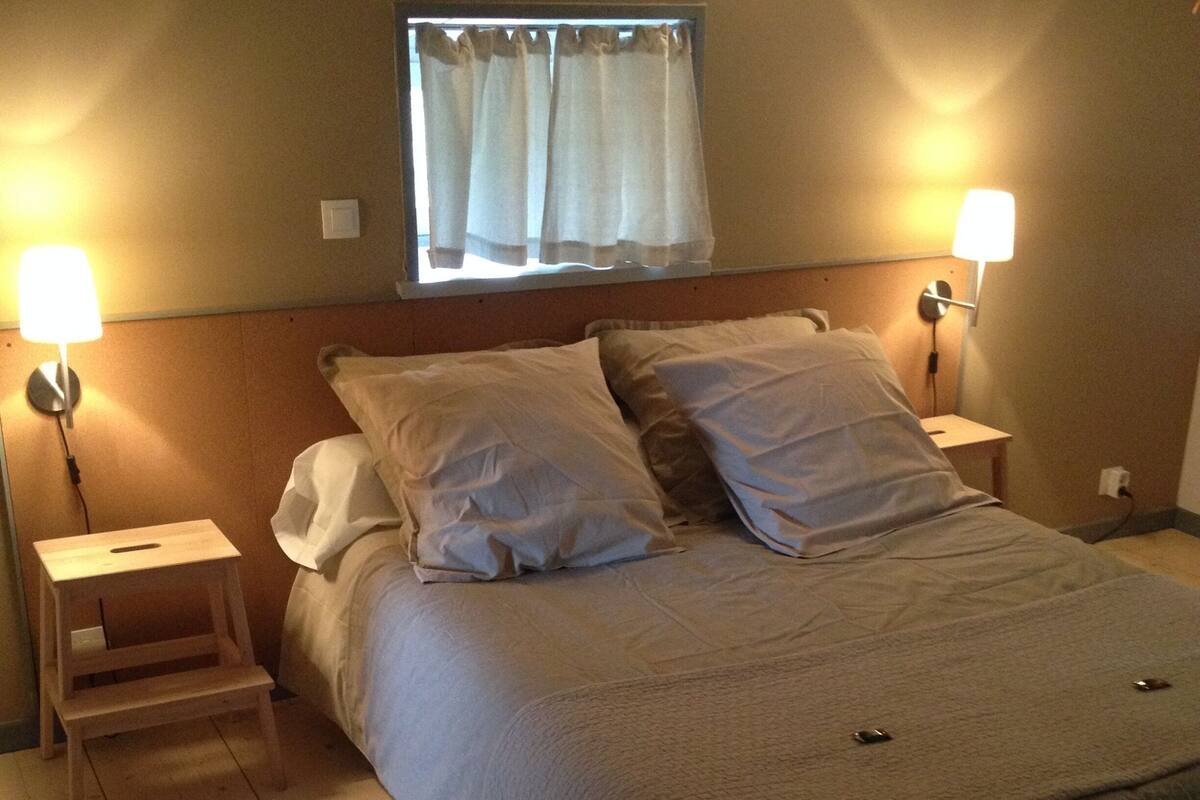 A bedroom with a bed, two bedside tables, a window with curtains, and two wall-mounted lamps.