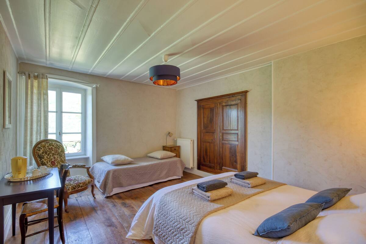 A bedroom with two beds, a wooden wardrobe, a table with a chair, and a window with curtains.