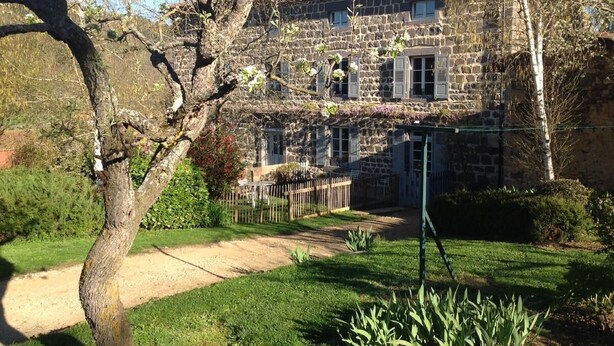 Un edificio de piedra con techo de tejas, patio cercado y un árbol en primer plano.