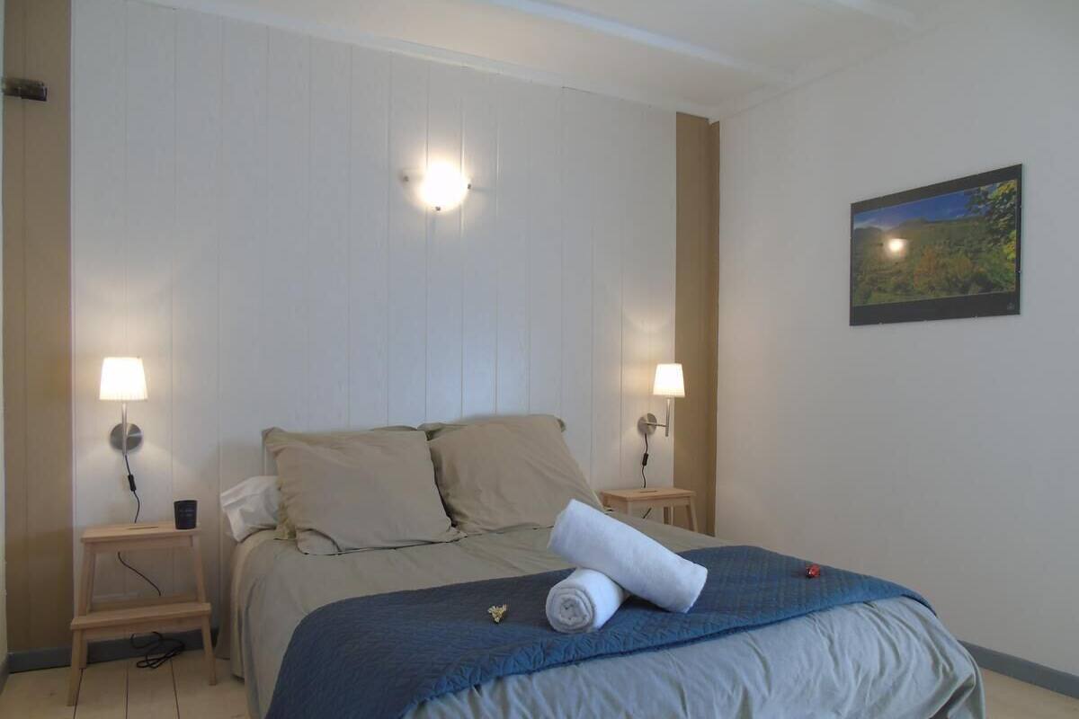 A neatly made bed with a blue and white duvet, two bedside lamps, a small wooden nightstand, and a framed landscape painting on the wall.