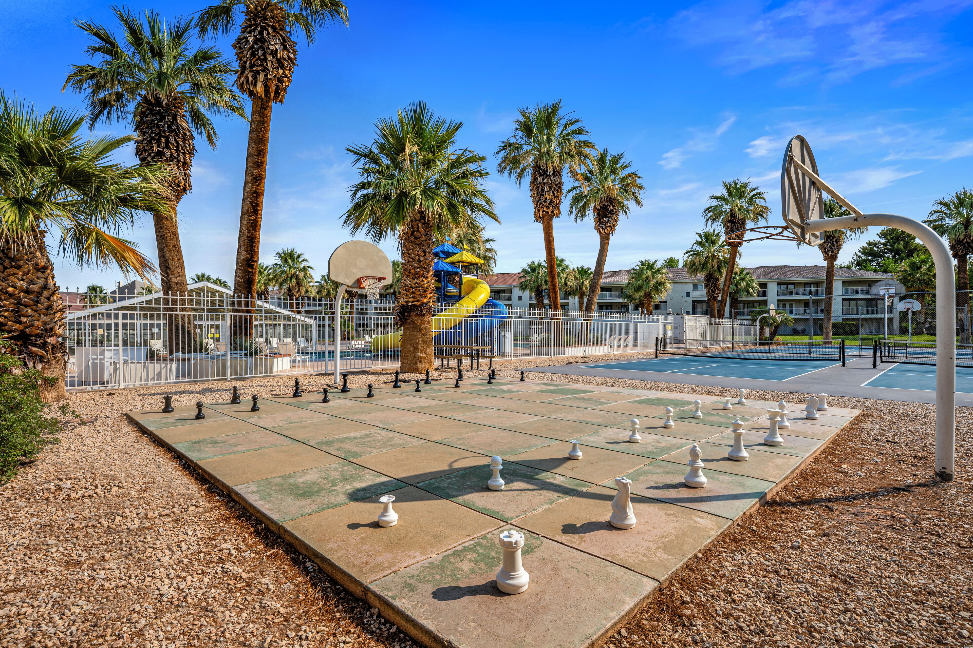 Ein Schachbrett auf einer Betonfläche, umgeben von Palmen und einem Basketballplatz.