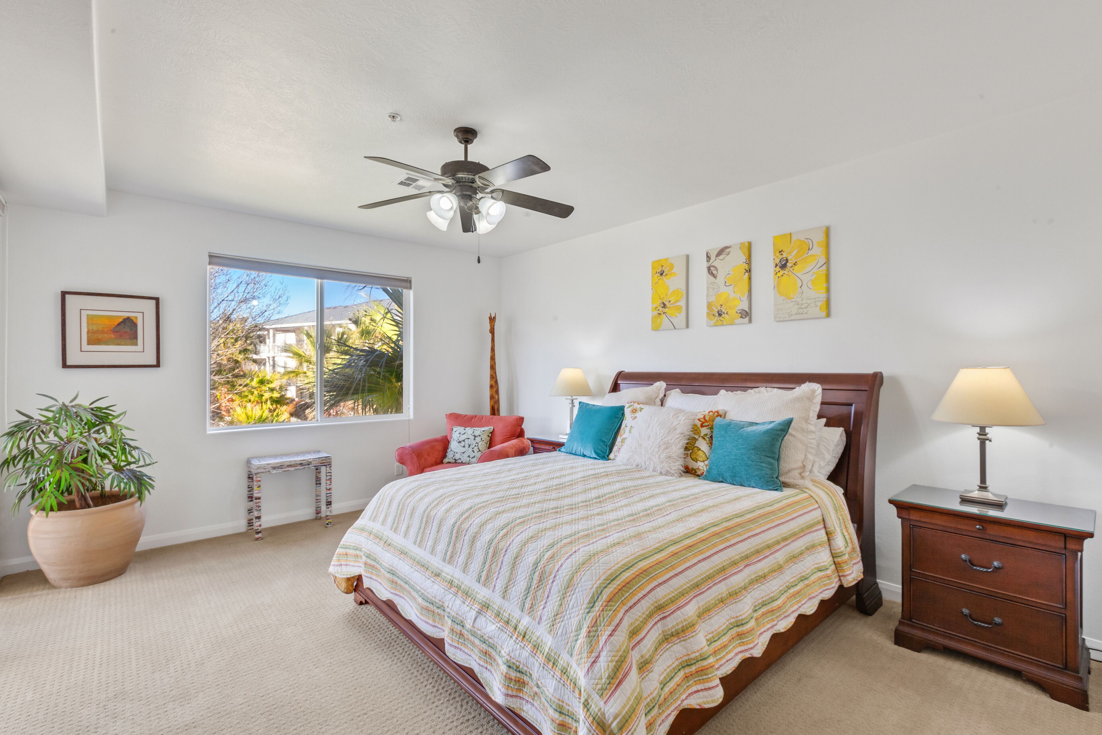 A bedroom with a bed, bedside table, nightstand, potted plant, window, and ceiling fan.