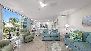 A spacious living area with a modern kitchen in the background, a large window with a view of palm trees, and a ceiling fan. - Island Beach Club 330B – Gulf view vacation rental condo on Sanibel Island (Sanibel)