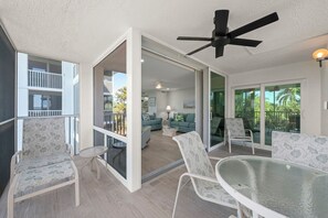 Un balcon avec une table et des chaises en verre, un ventilateur de plafond et une vue sur un immeuble doté de balcons.