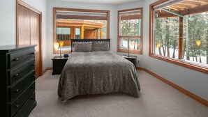 Un dormitorio con una cama grande, cabecera de madera, dos mesitas de noche, un aparador y ventanas amplias que ofrecen vistas a una terraza de madera y árboles.