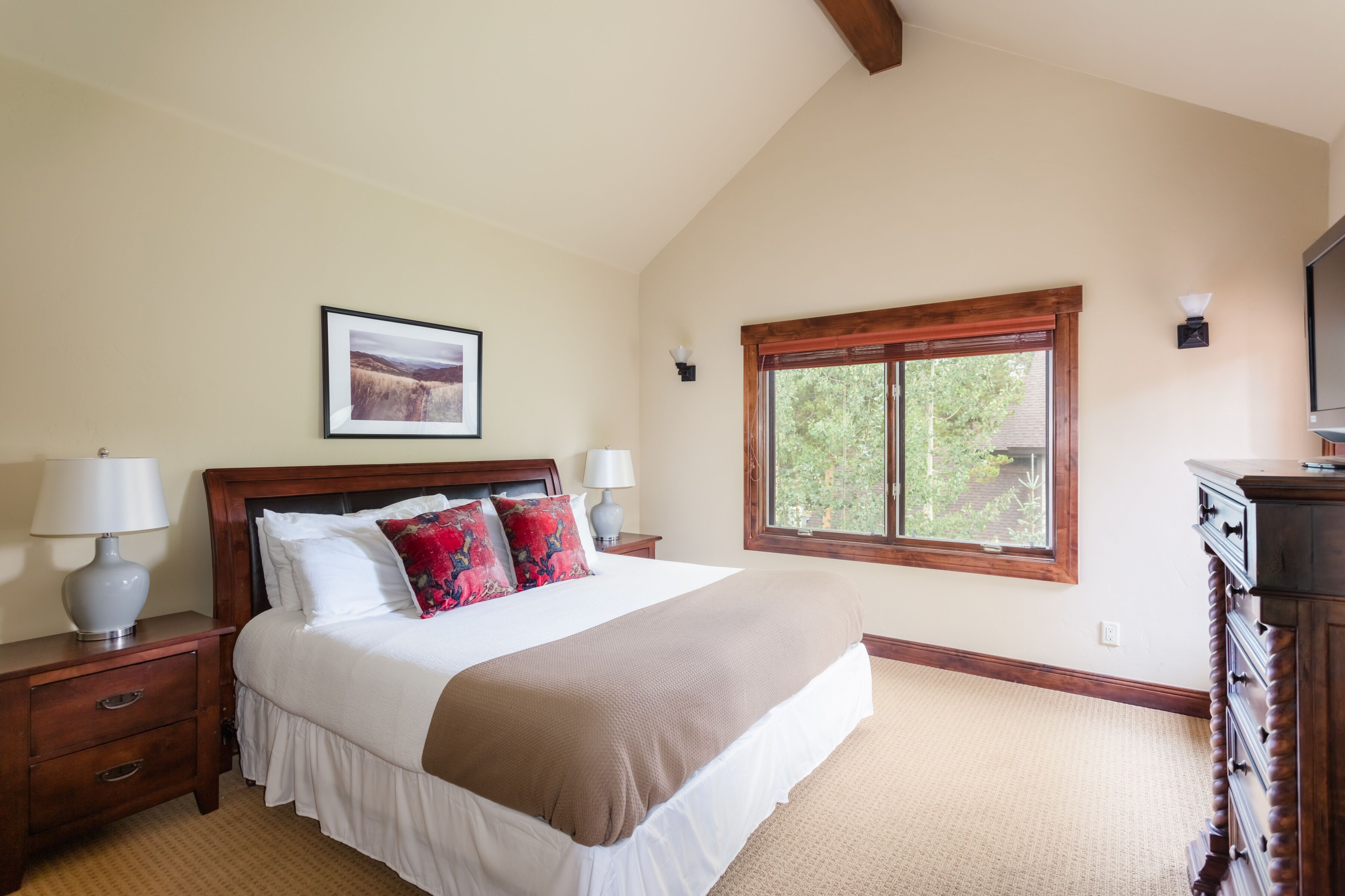 A bedroom with a large bed, wooden headboard, two bedside tables with lamps, a dresser, a window with a view, and a framed picture on the wall.