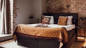 A bedroom with a brick wall, a bed with a wooden headboard, a nightstand, and a leather armchair. - Broederenklooster (Zutphen)