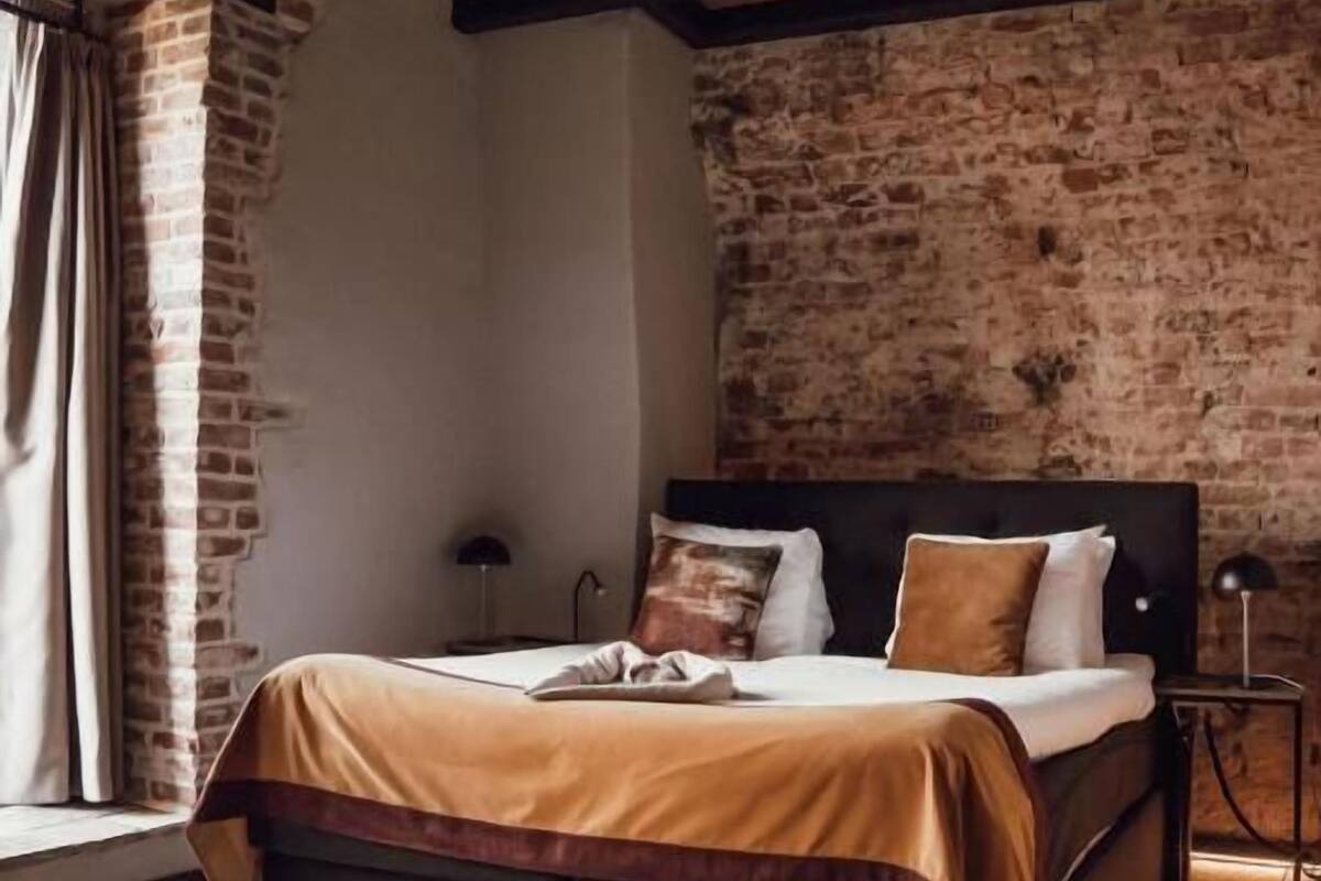 A bedroom with a brick wall, a bed with a wooden headboard, a nightstand, and a leather armchair.