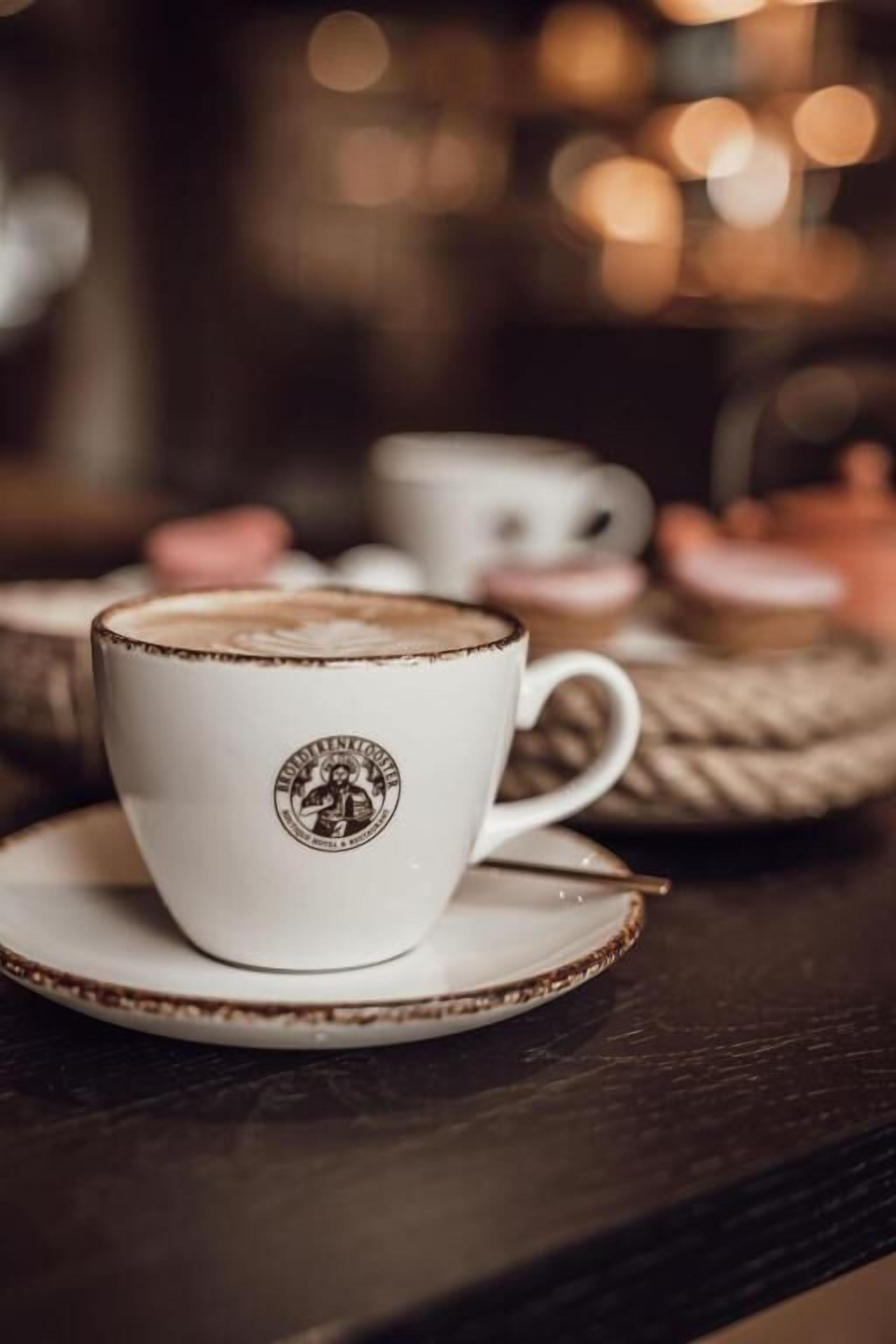 A white cup with a logo on a saucer, placed on a wooden table with other cups and pastries in the background.