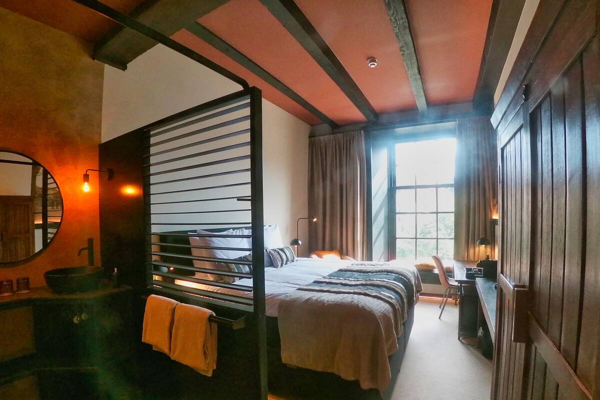 A bedroom with a bed, a desk, a chair, a window, and a bathroom with a sink and mirror.