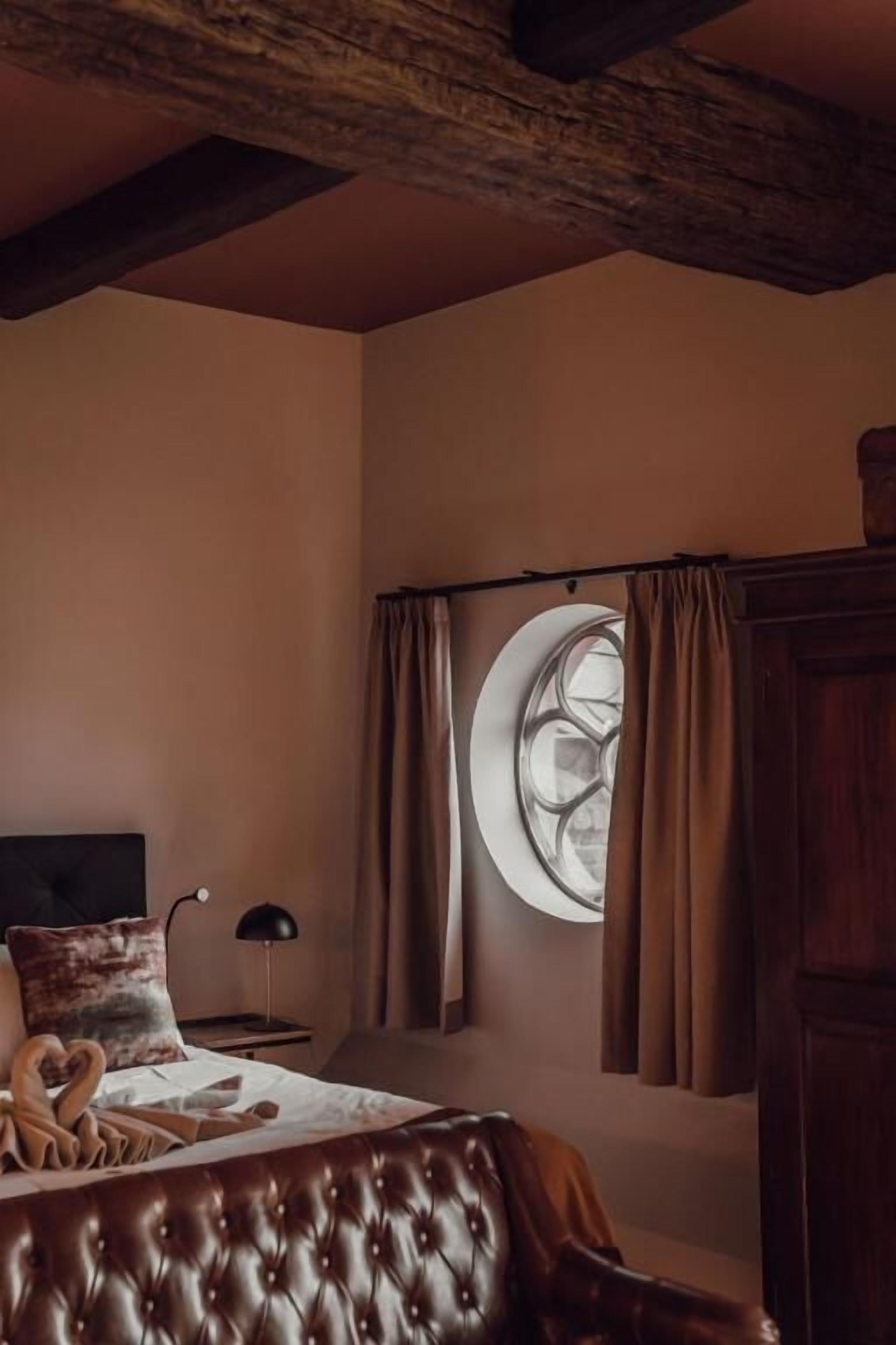 A bedroom with a tufted headboard, a round window with curtains, a bed with a heart-shaped pillow, and a nightstand with a lamp.