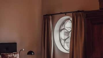 A bedroom with a tufted headboard, a round window with curtains, a bed with a heart-shaped pillow, and a nightstand with a lamp.