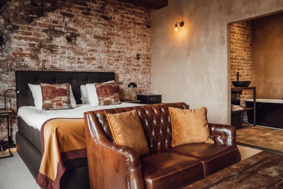 A bedroom with a brick wall, a bed with a black headboard, a leather sofa, and a small table with a lamp.