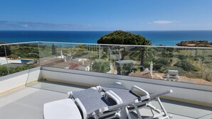 Un balcone con una poltrona in vimini bianco e un tavolino, affacciato su un paesaggio costiero con vista libera sul mare e colline lontane.