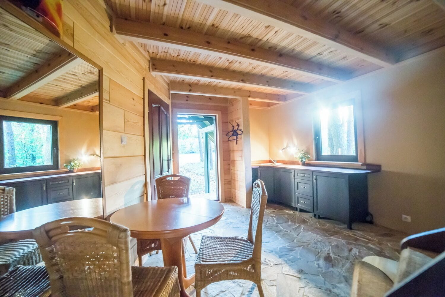 A wooden interior with a round table and chairs, a kitchen area with dark cabinets, and a stone floor.