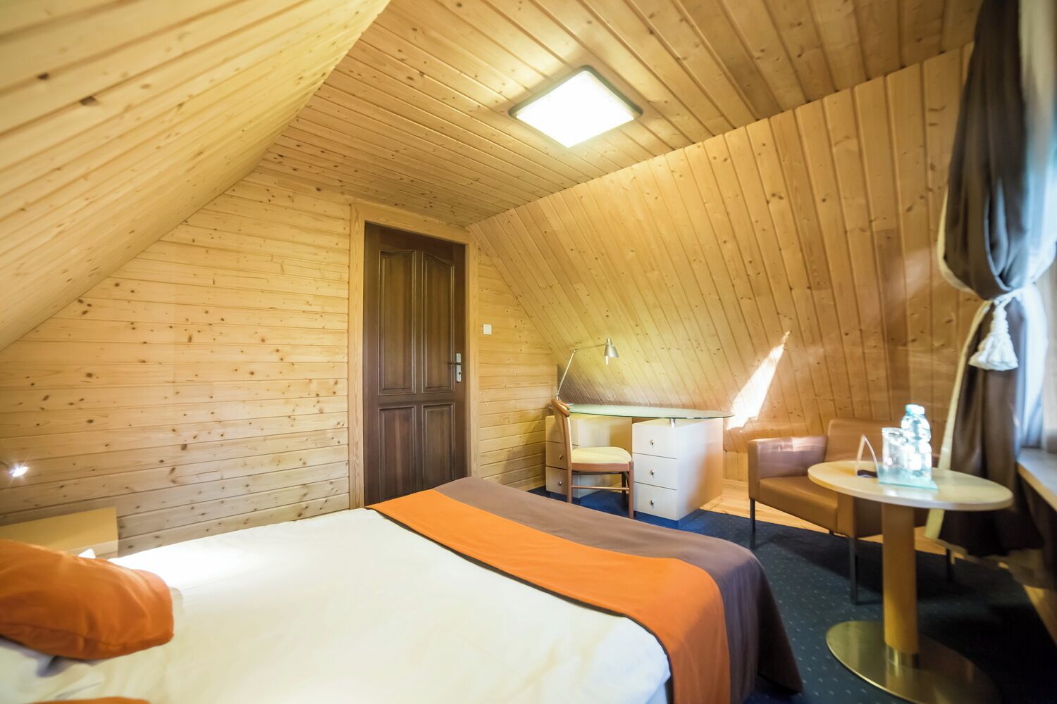 Une chambre mansardée chaleureuse, avec un lit, un bureau, une chaise et une petite table sur laquelle se trouve une lampe.