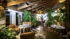 An indoor dining area with wicker furniture, round tables, and potted plants.
