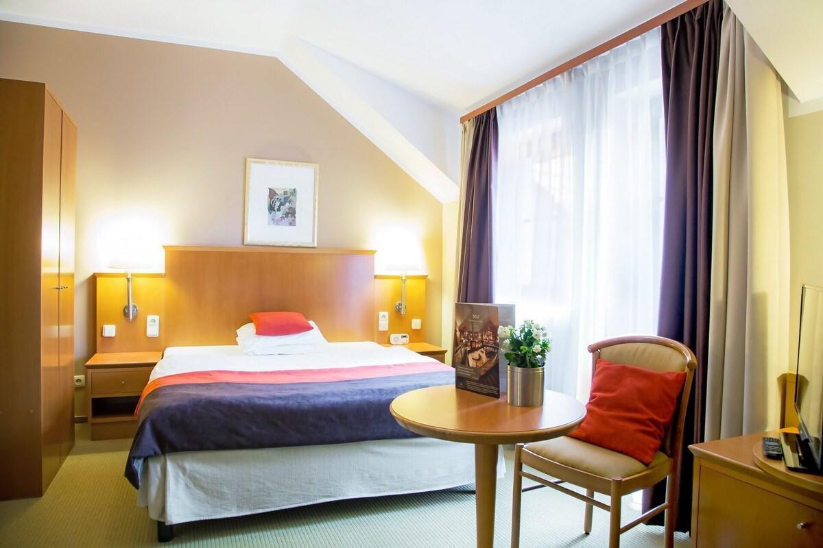 Quarto de hotel com cama, mesas de cabeceira, escrivaninha, cadeira e janela com cortinas.