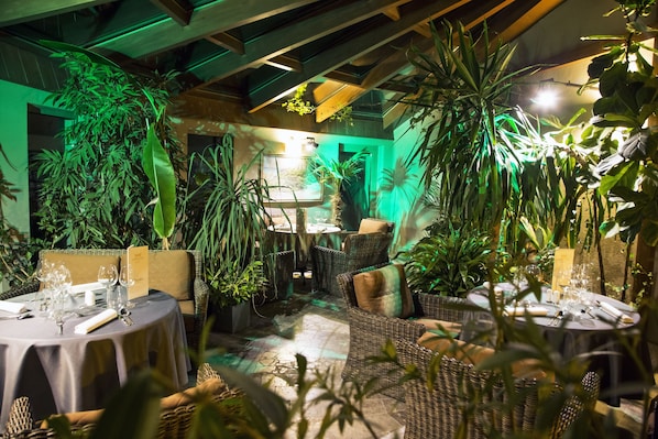 A restaurant with wicker furniture, tables set with glassware, and lush greenery.