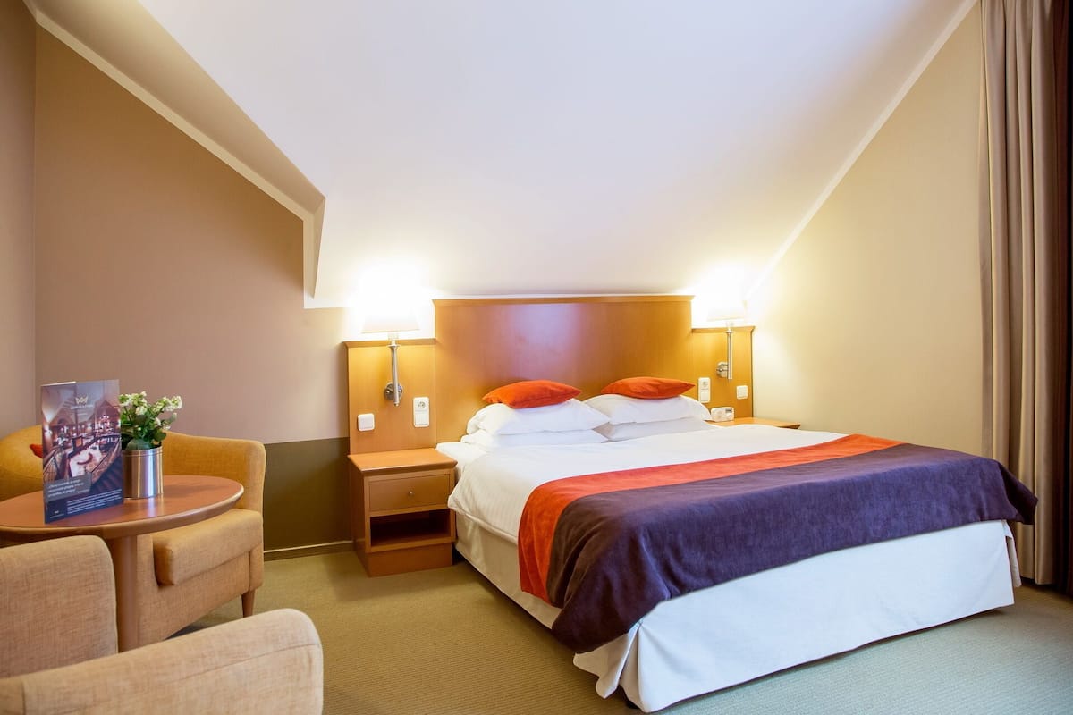 A hotel room with a bed, two bedside tables, a chair, and a small table with a vase and a brochure.