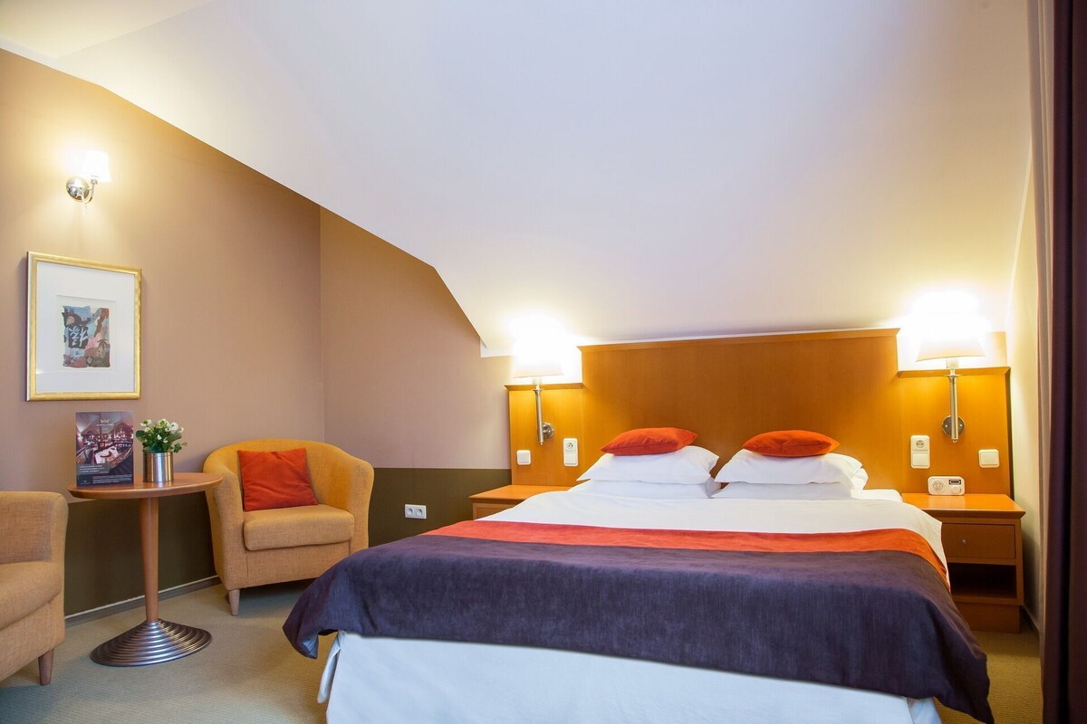 Quarto de hotel com uma cama, duas mesas de cabeceira, uma cadeira, uma pequena mesa com uma planta e duas luminárias na parede.