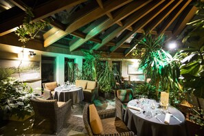 An indoor dining area with wicker furniture, tables set for dining, and green plants.