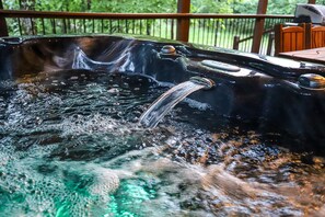 A hot tub with a waterfall feature, surrounded by a wooden deck and outdoor furniture. - Lazy Pine Lodge: 4BR, 4BA, Sleeps 14 (Broken Bow)