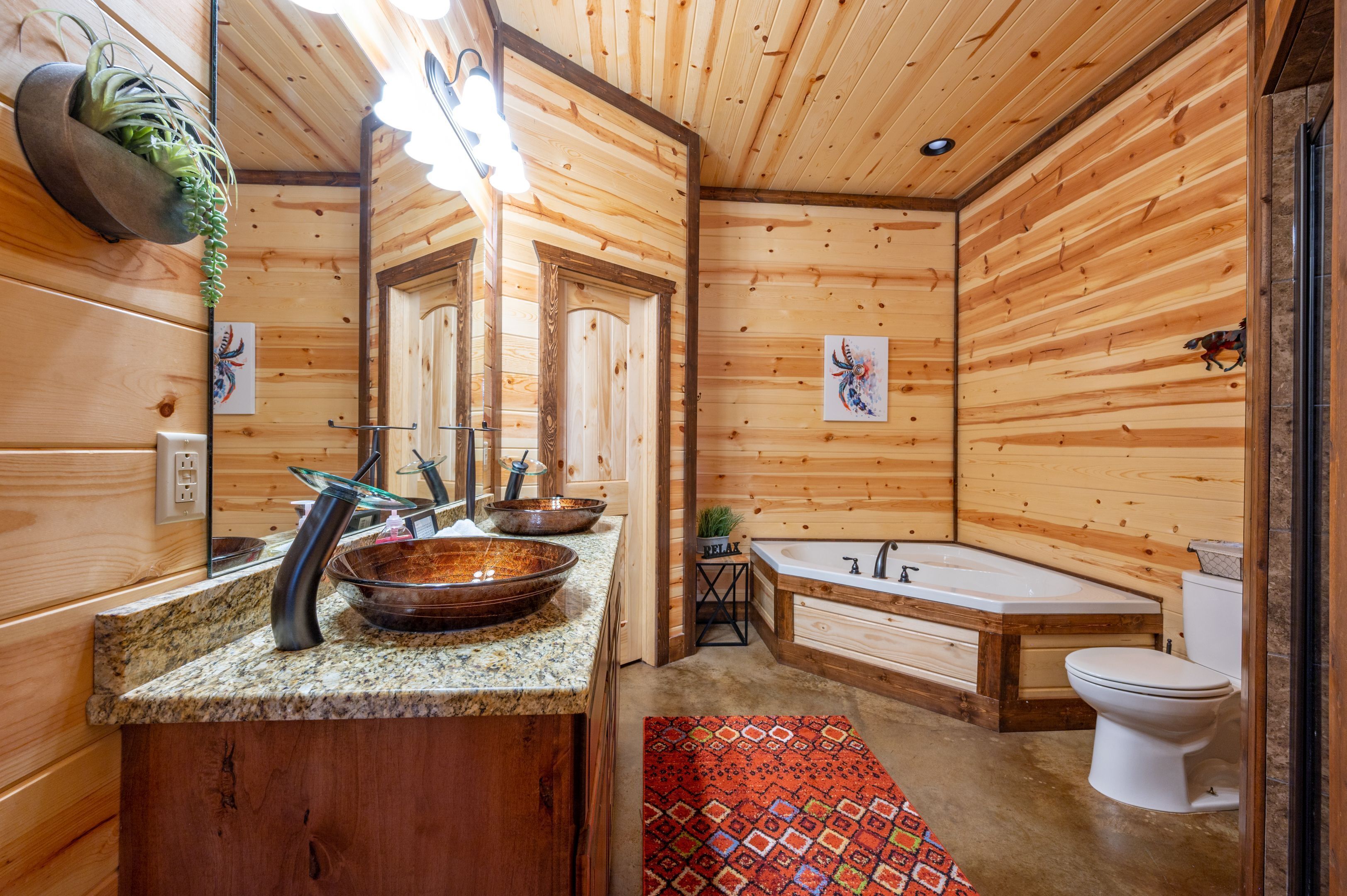 Baño de cabaña de madera con encimera de granito, un espejo grande, una bañera y un inodoro.