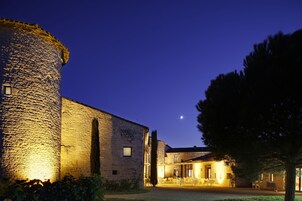 Un edificio de piedra con una torre cilíndrica, un patio con mesas y sillas, y un árbol.
