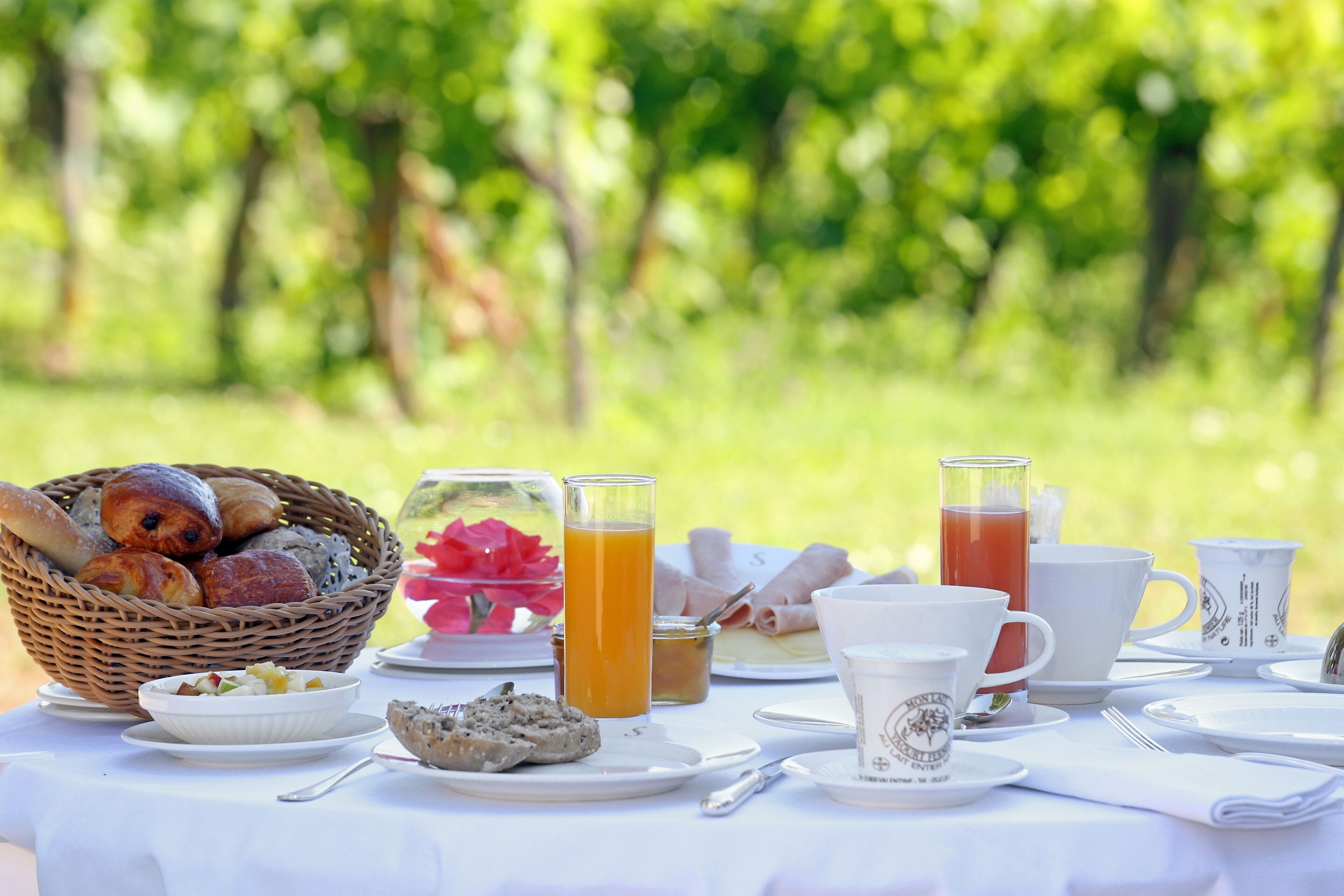 A breakfast table set outdoors with a basket of bread, a bowl of fruit, a glass of orange juice, and several cups and saucers.