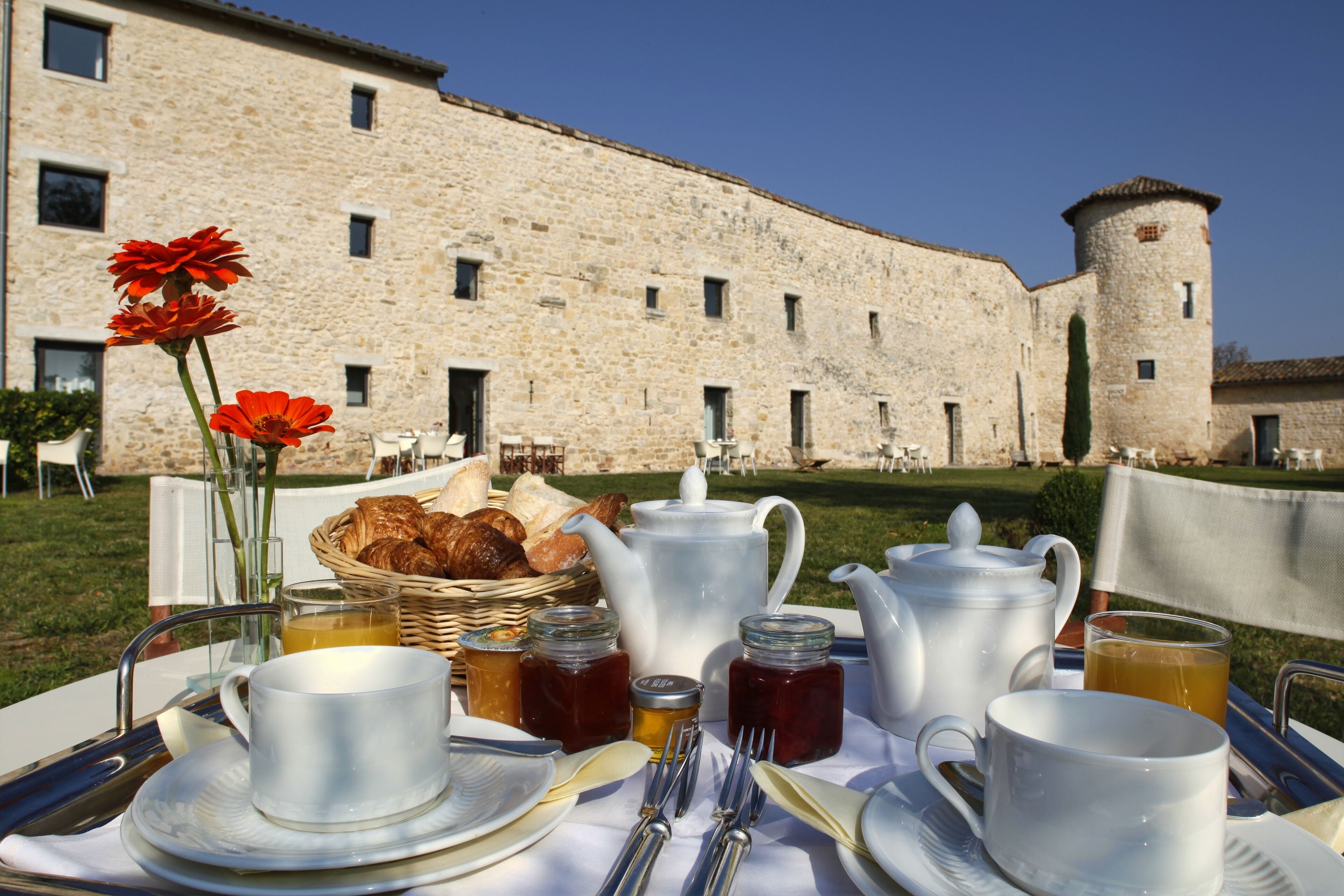 A breakfast setup with a table, teapot, cups, and pastries in front of a stone building with a tower.