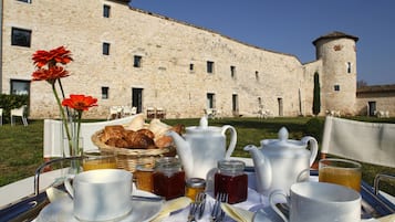 A breakfast setup with a table, teapot, cups, and pastries in front of a stone building with a tower.
