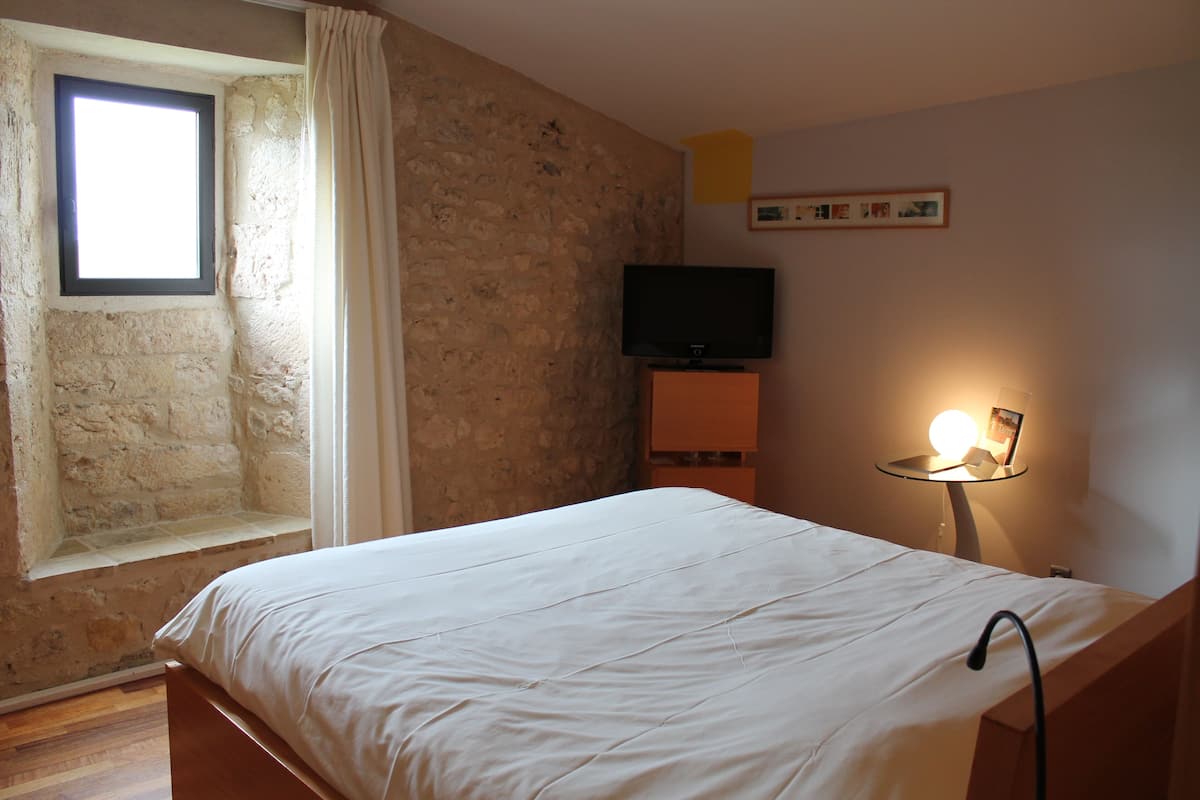 Une chambre avec un mur en pierre, une fenêtre avec des rideaux, un lit avec une literie blanche, un téléviseur posé sur un meuble et une table de chevet avec une lampe et un cadre photo.