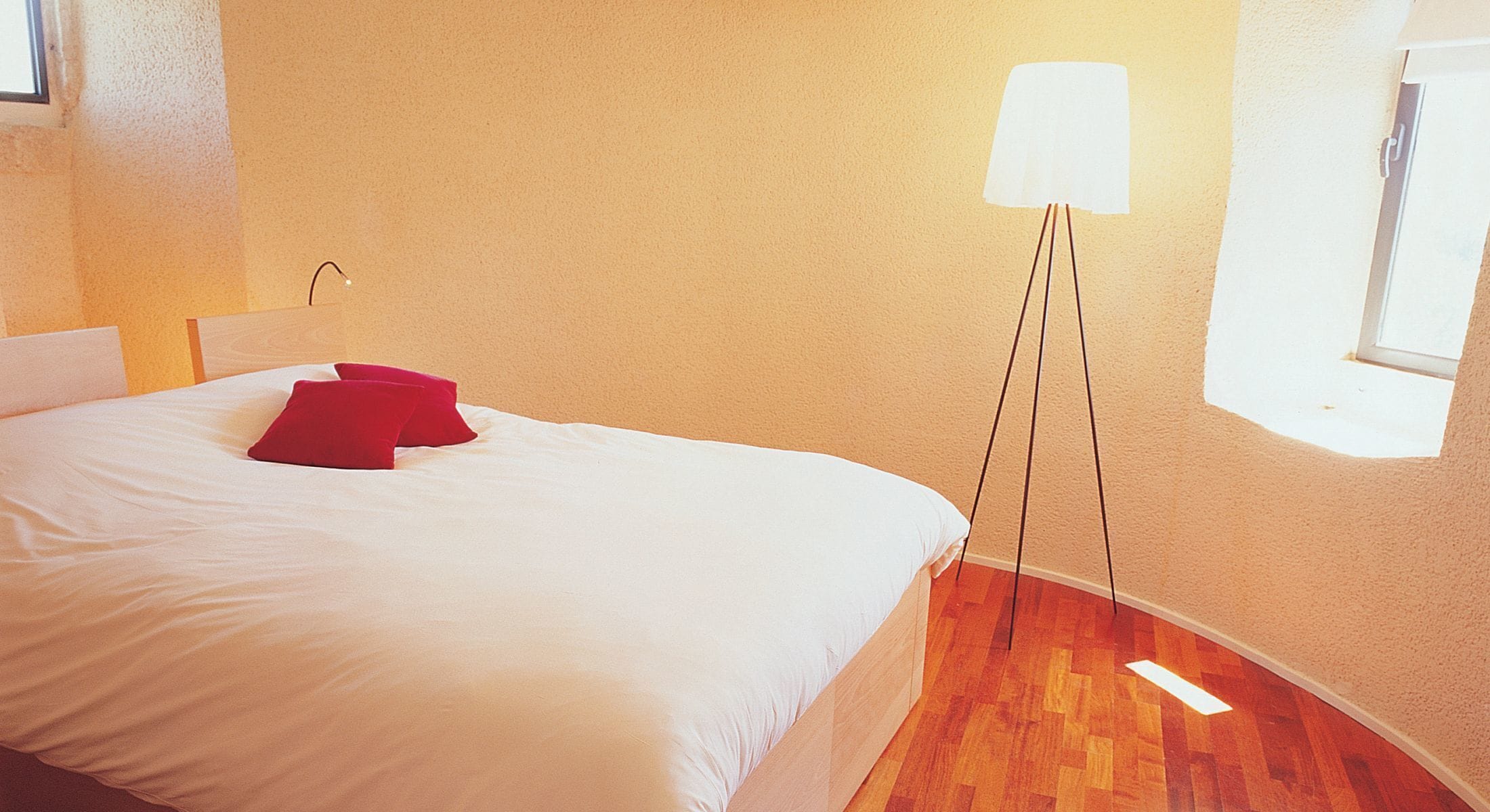 Uma cama bem arrumada com almofadas brancas e vermelhas, um abajur de chão e uma janela que oferece luz natural.