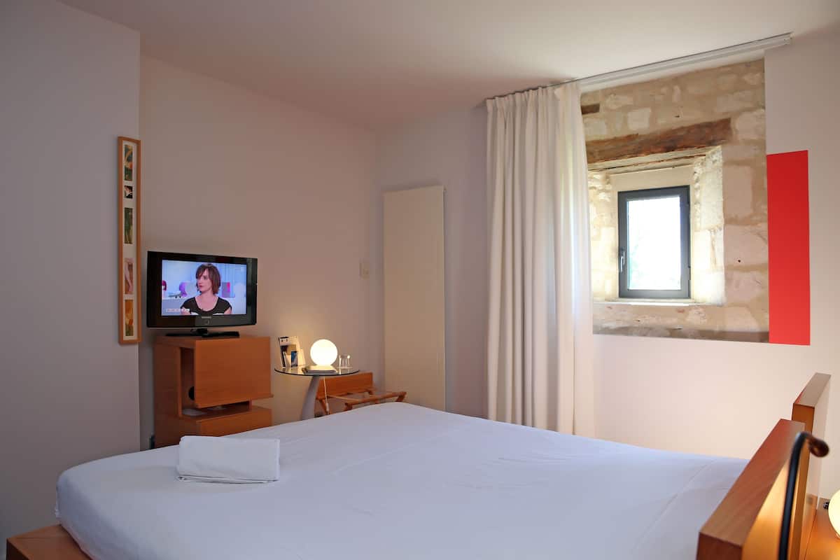 Une chambre d’hôtel avec un lit, un téléviseur posé sur un meuble en bois, une petite table avec une lampe et une fenêtre avec des rideaux.