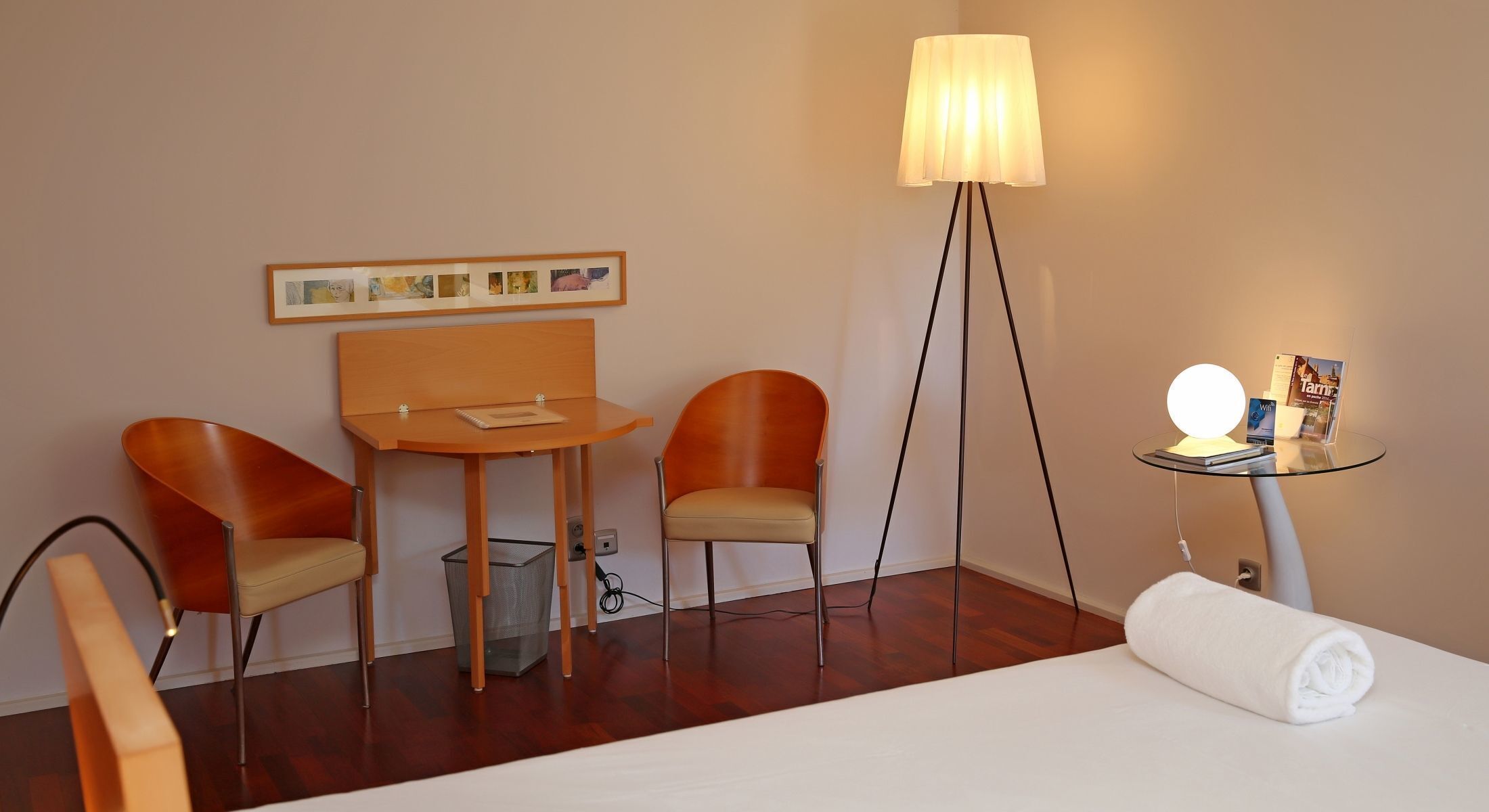 A hotel room with a bed, two chairs, a small table, a lamp, and a wall-mounted picture frame.