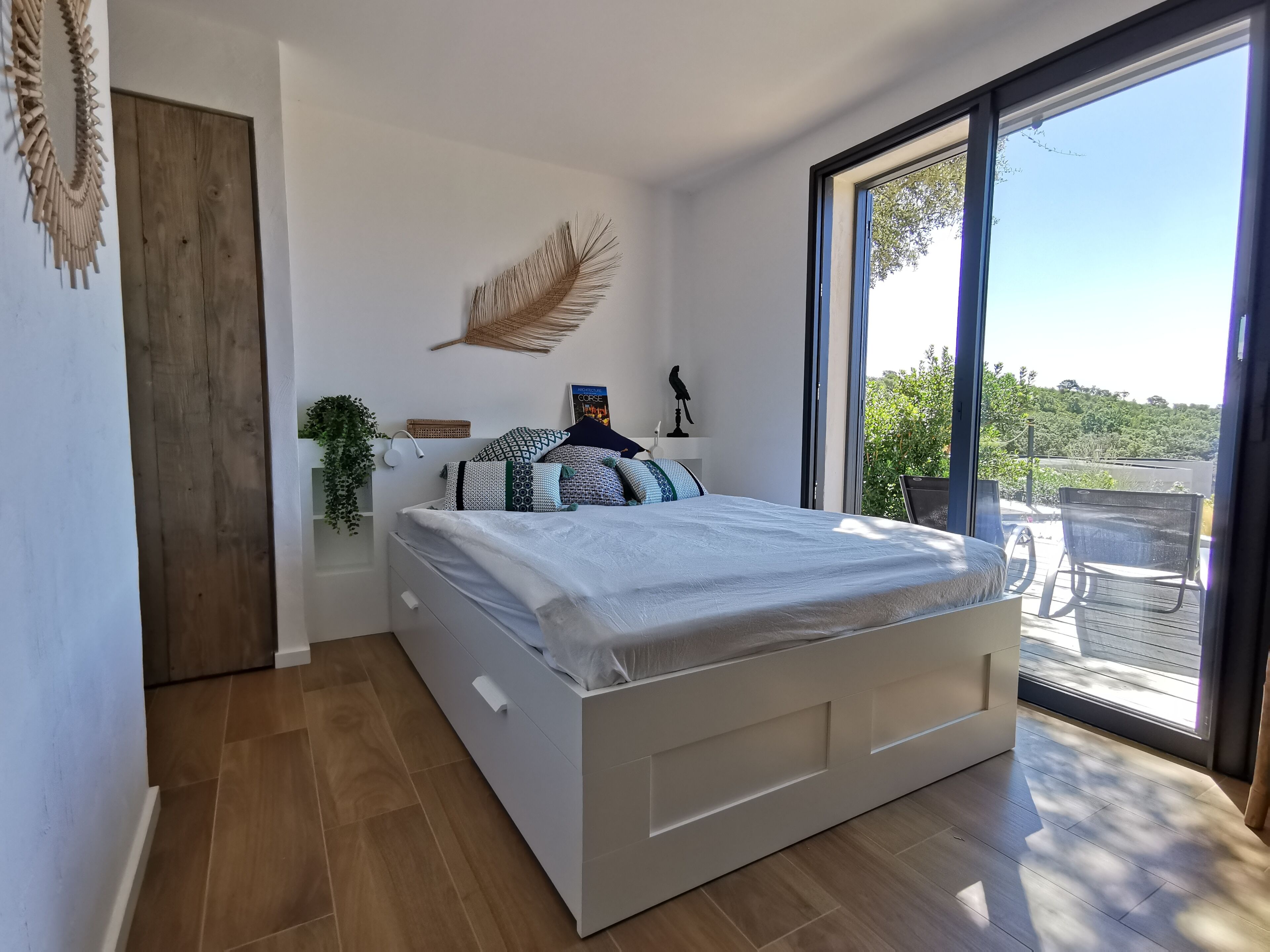 A modern bedroom with a large bed, a wooden door, a view of the outdoors, and a ceiling fan.