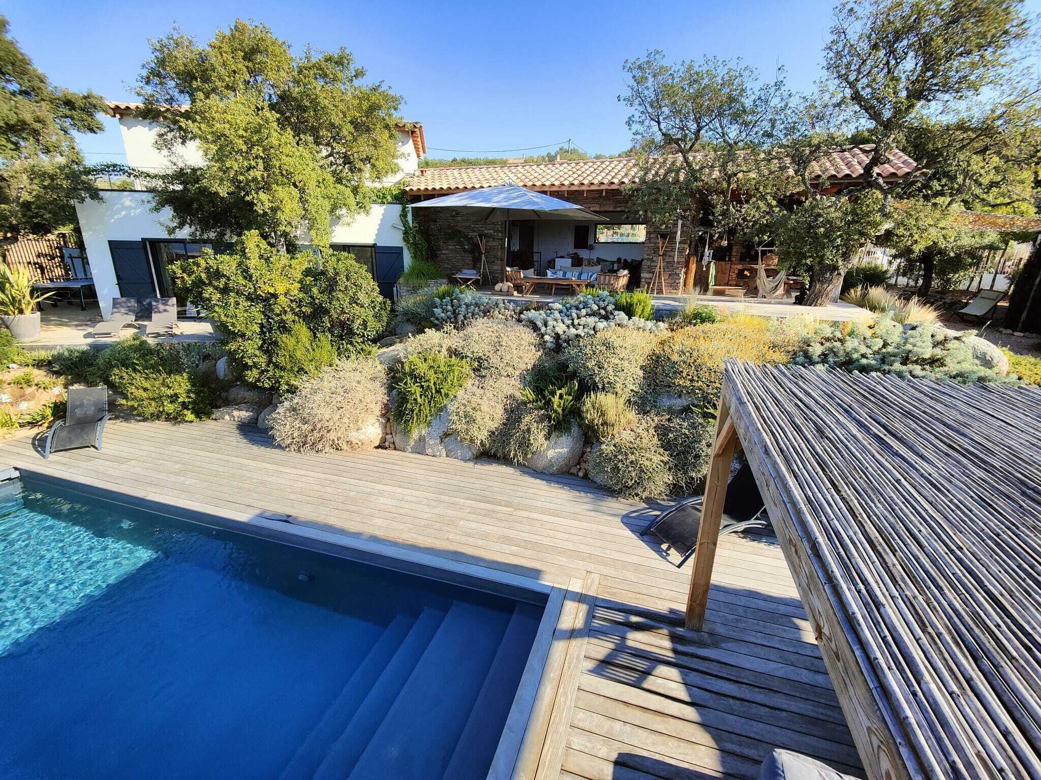 A pool area with a wooden deck, lounge chairs, and a stone wall with plants.