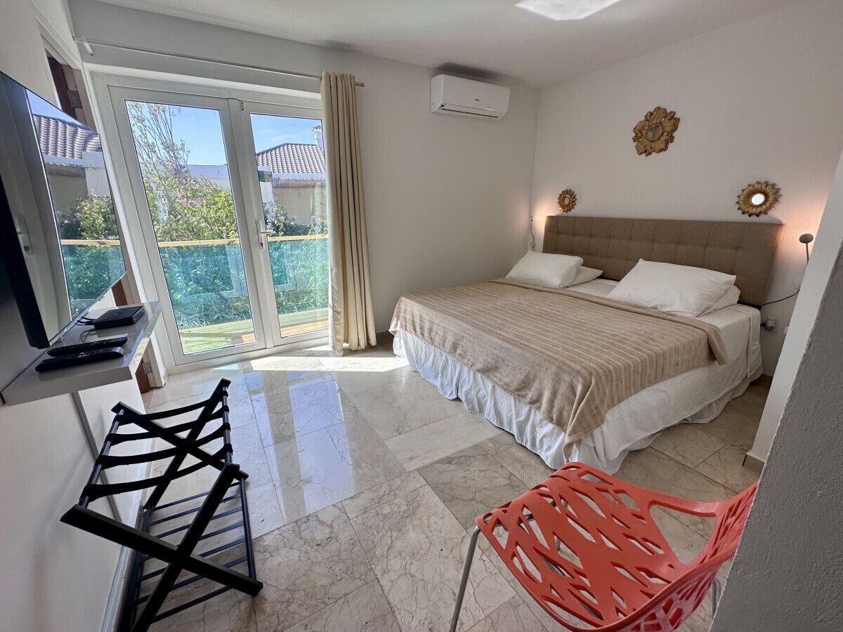 Un dormitorio con cama, televisor, tendedero, silla y balcón con cortinas.