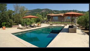 Une piscine entourée de chaises longues et de parasols, bordée de verdure et avec une chaîne de montagnes en toile de fond.