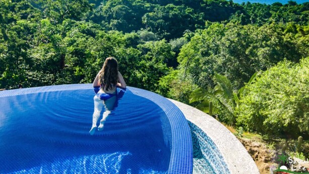 Una persona sentada al borde de una piscina con vistas a un bosque y al océano.