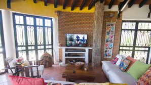 Una sala de estar con pared de ladrillo, un televisor sobre un soporte blanco, un sofá con cojines de colores y una mesa de centro de madera.